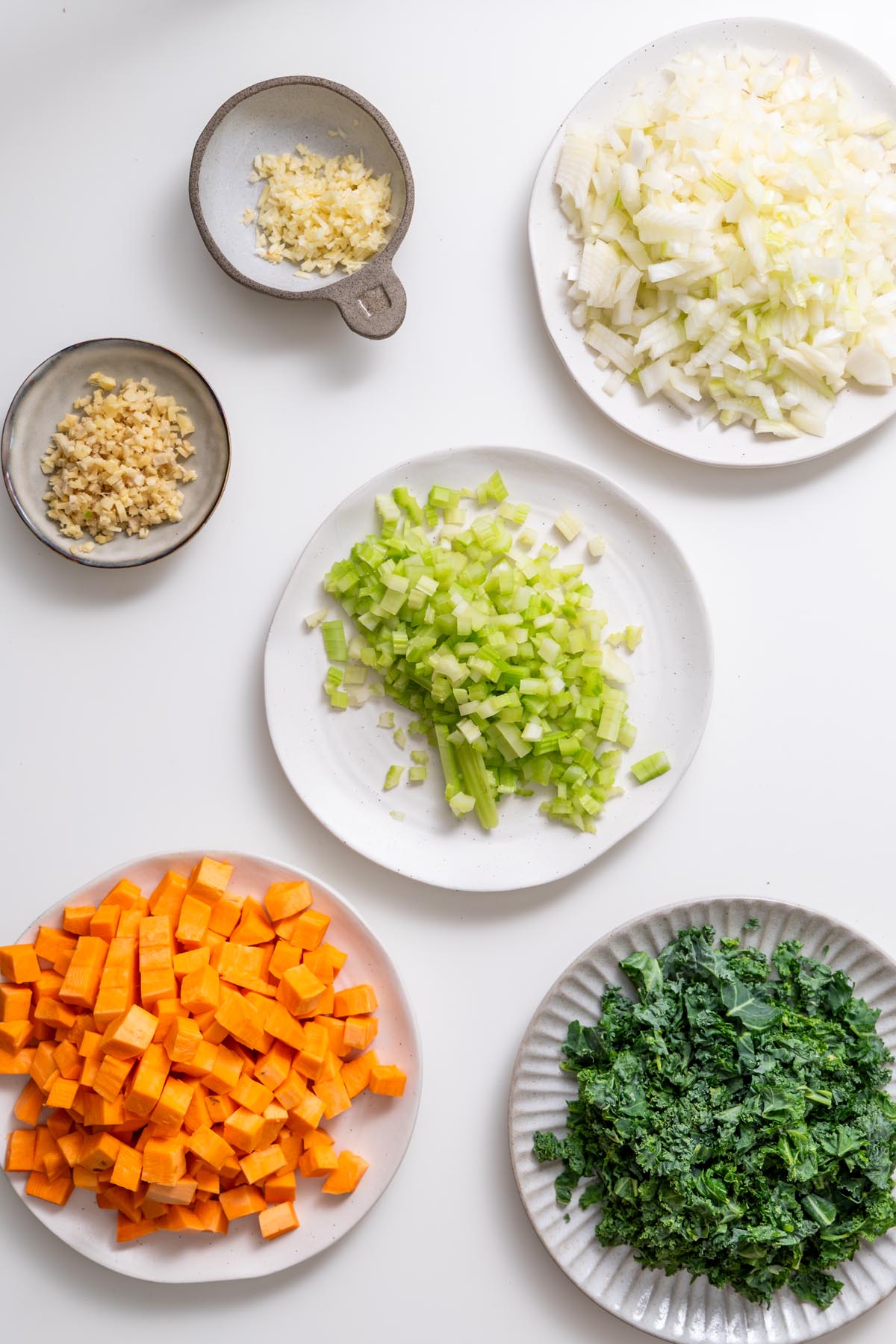 Chopped sweet potato, kale, celery, onion, garlic, and ginger prepped on plates for red lentil soup.