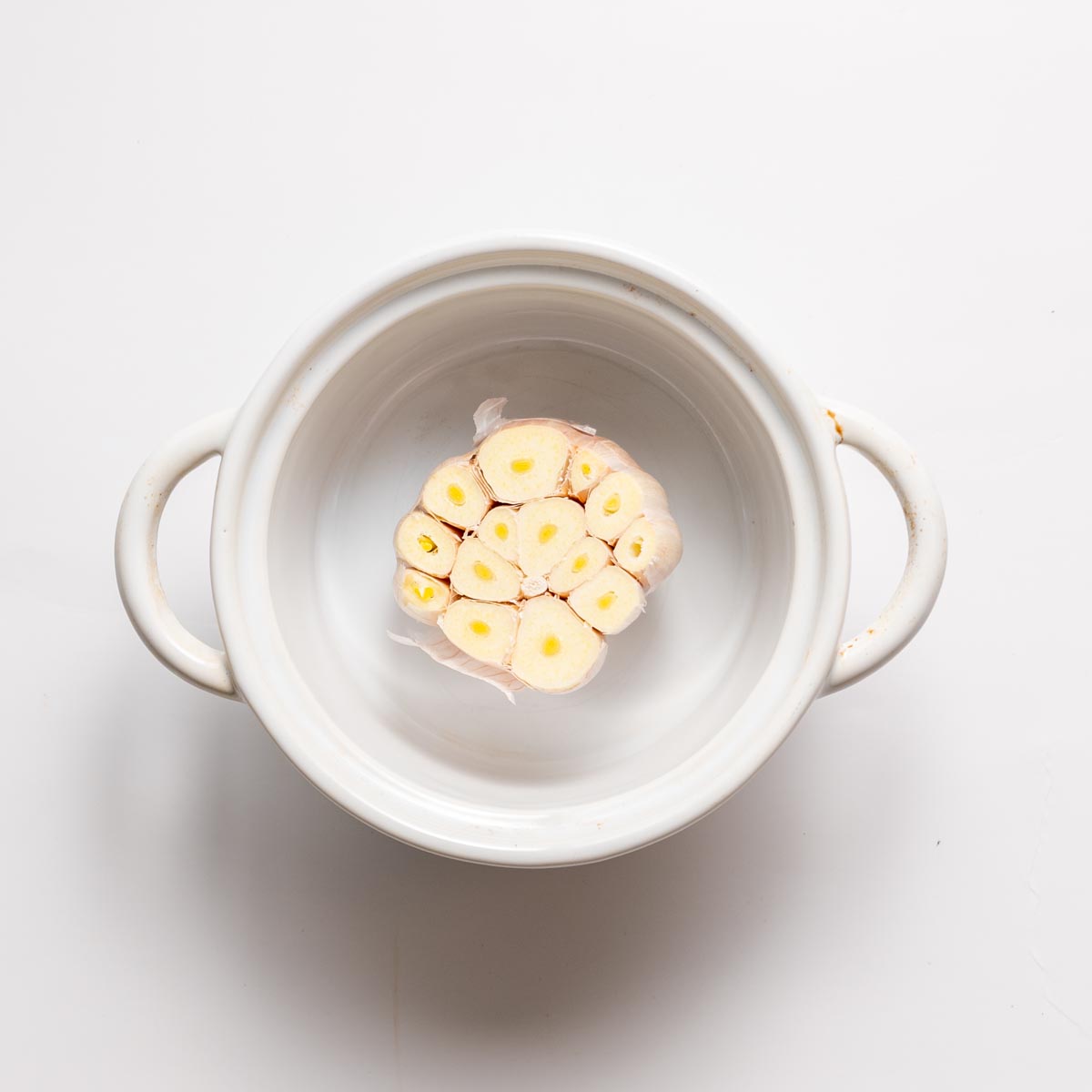 Raw garlic bulb sliced across the top in a small white baking dish ready for roasting.