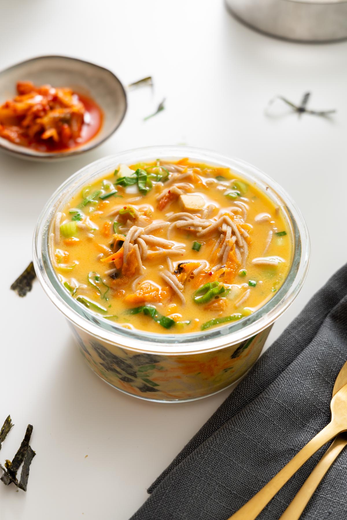 Close-up of miso noodle soup in a meal prep container with kimchi on the side.