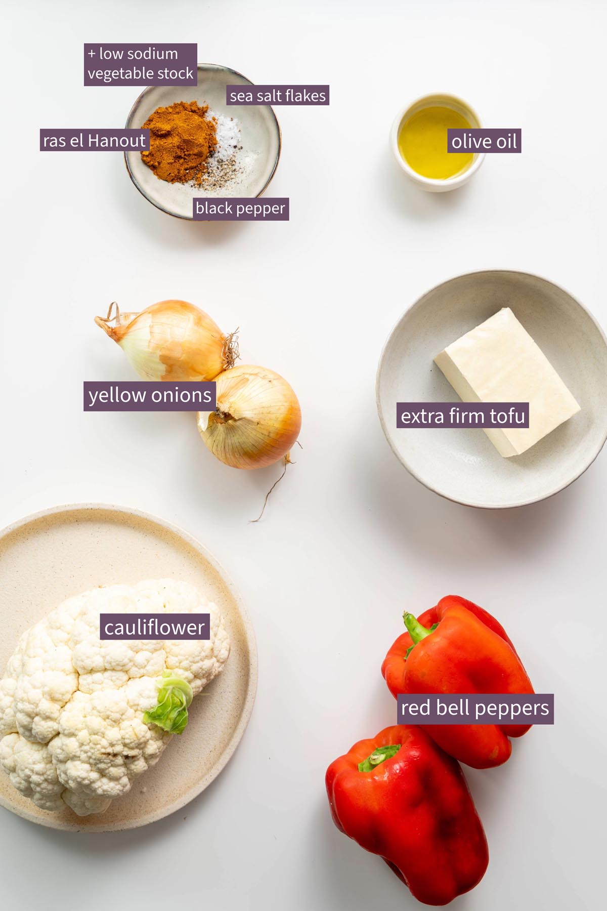 Vibrant ingredients for roasted cauliflower and red bell pepper soup on a white surface, including cauliflower, red bell peppers, yellow onions, tofu, and spices.