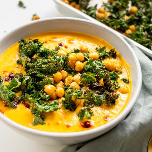 Close-up of creamy roasted cauliflower and red bell pepper soup topped with crispy kale and chickpeas in a white bowl.