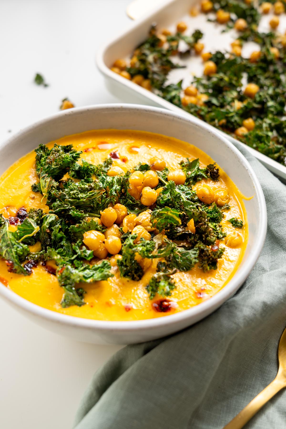 Close-up of creamy roasted cauliflower and red bell pepper soup topped with crispy kale and chickpeas in a white bowl.