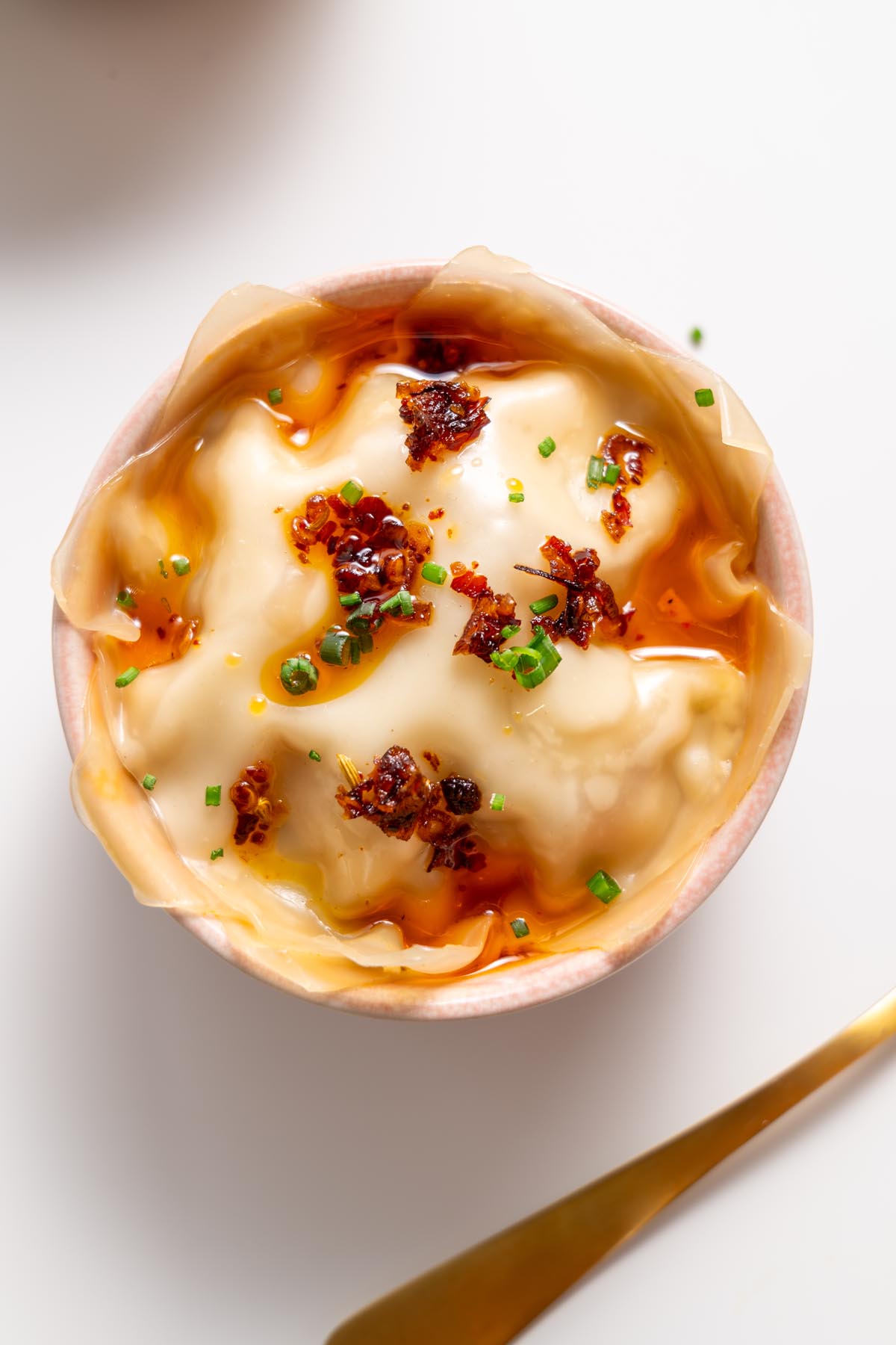 Nhìn từ trên cao của món lasagna súp bánh bao trong một chiếc bát màu hồng phủ dầu ớt và hành lá, với một chiếc thìa vàng bên cạnh