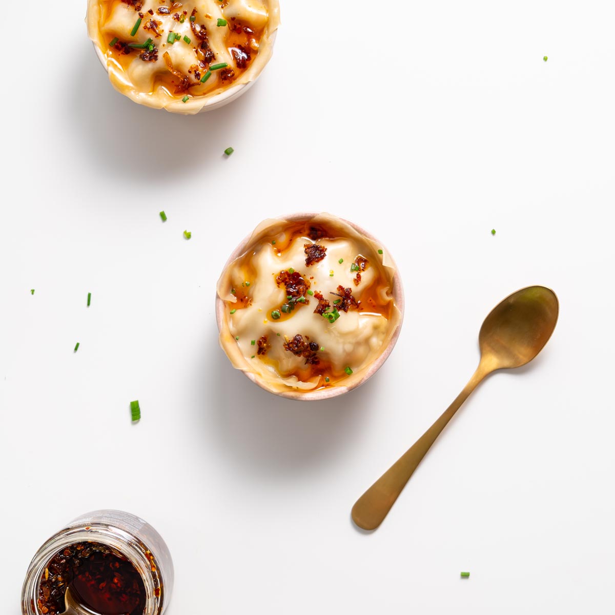 Hai tô lasagna bánh bao phủ dầu ớt giòn và hành lá, kèm lọ dầu ớt và thìa vàng trên mặt trắng