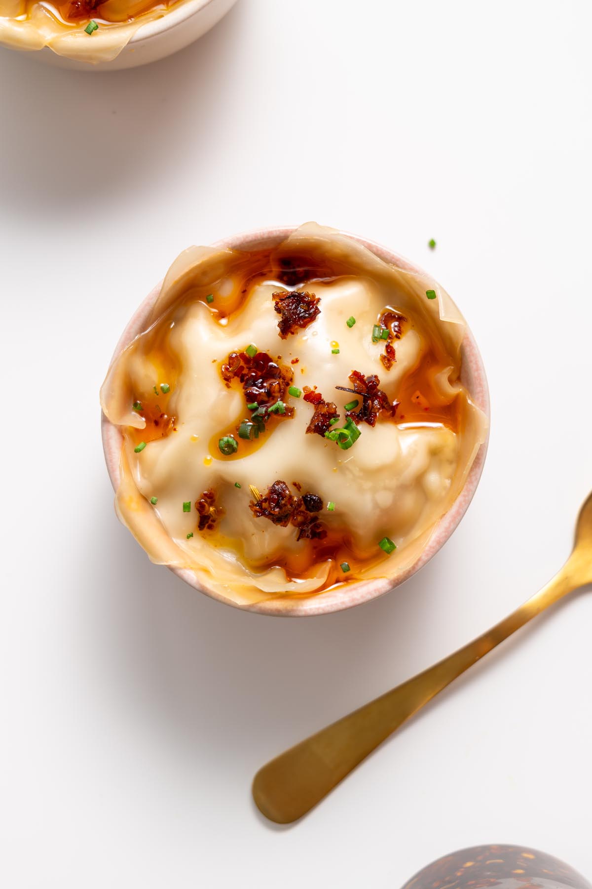 Nhìn từ trên cao của món lasagna súp bánh bao trong một chiếc bát màu trắng rưới dầu ớt và trang trí bằng hành lá, với một chiếc thìa vàng bên cạnh