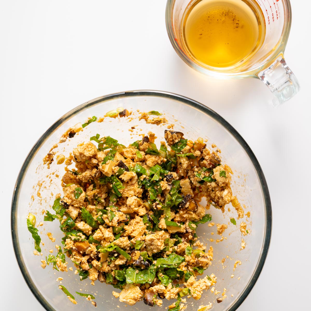 Đậu hủ trộn với nấm đông cô, bắp cải và hành lá đựng trong tô màu trắng, bên cạnh có một cốc đong nước dùng