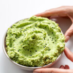 Protein edamame avocado spread in a white bowl, held by Sarah Cobacho on a white background.