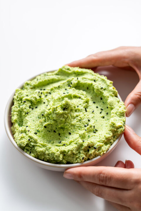 Protein edamame avocado spread in a white bowl, held by Sarah Cobacho on a white background.