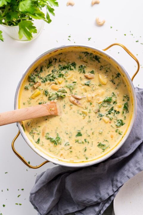 Overhead view of creamy vegan Zuppa Toscana with white beans, kale, and potatoes in a large pot with wooden spoon.