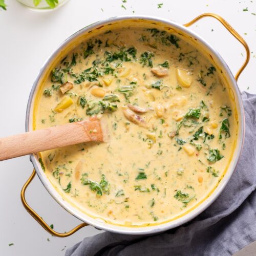 Overhead view of creamy vegan Zuppa Toscana with white beans, kale, and potatoes in a large pot with wooden spoon.