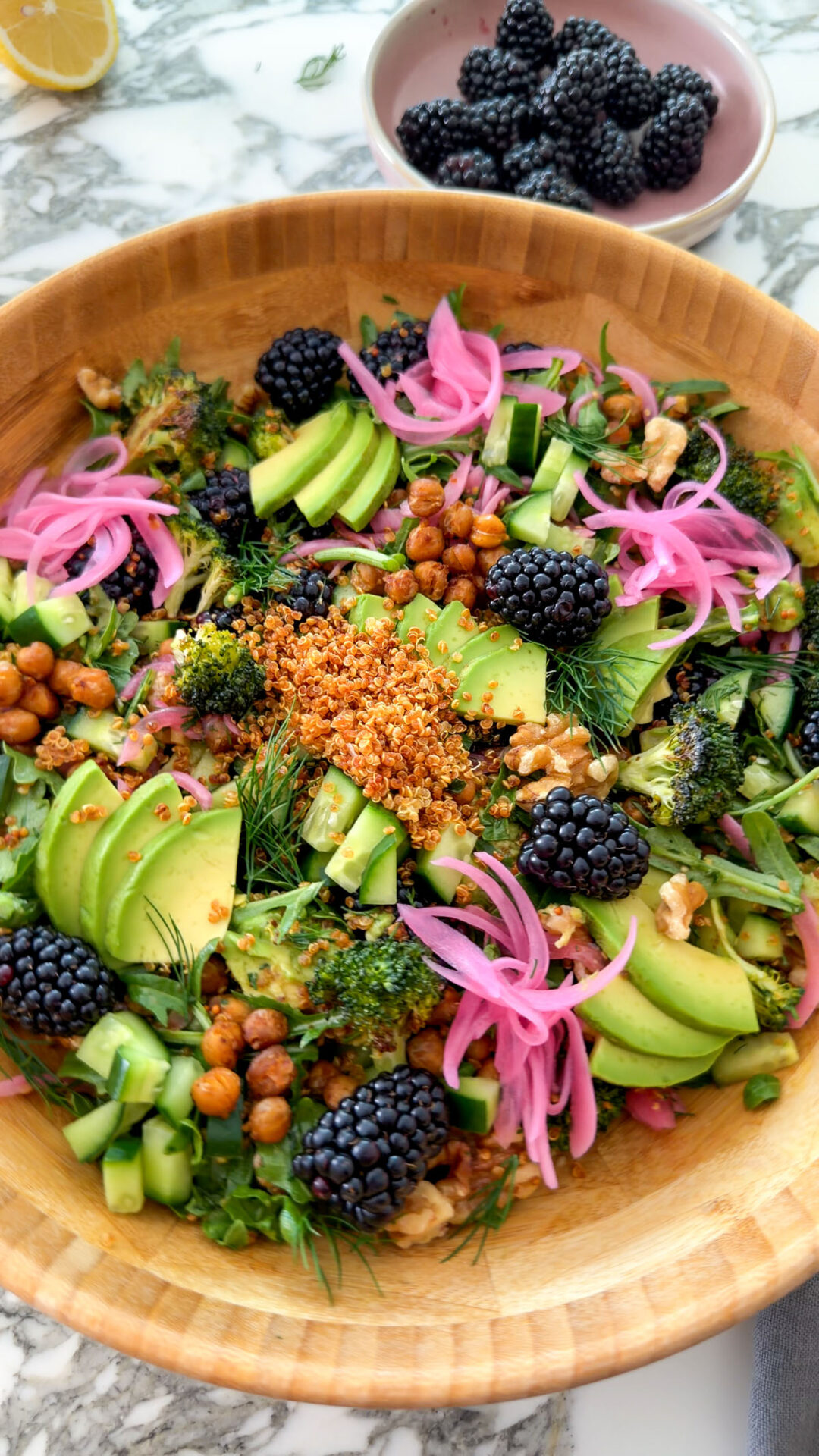 Close-up of crispy quinoa and blackberry salad with avocado, cucumber, roasted broccoli, chickpeas, dill, walnuts, and pickled onions in a wooden bowl.