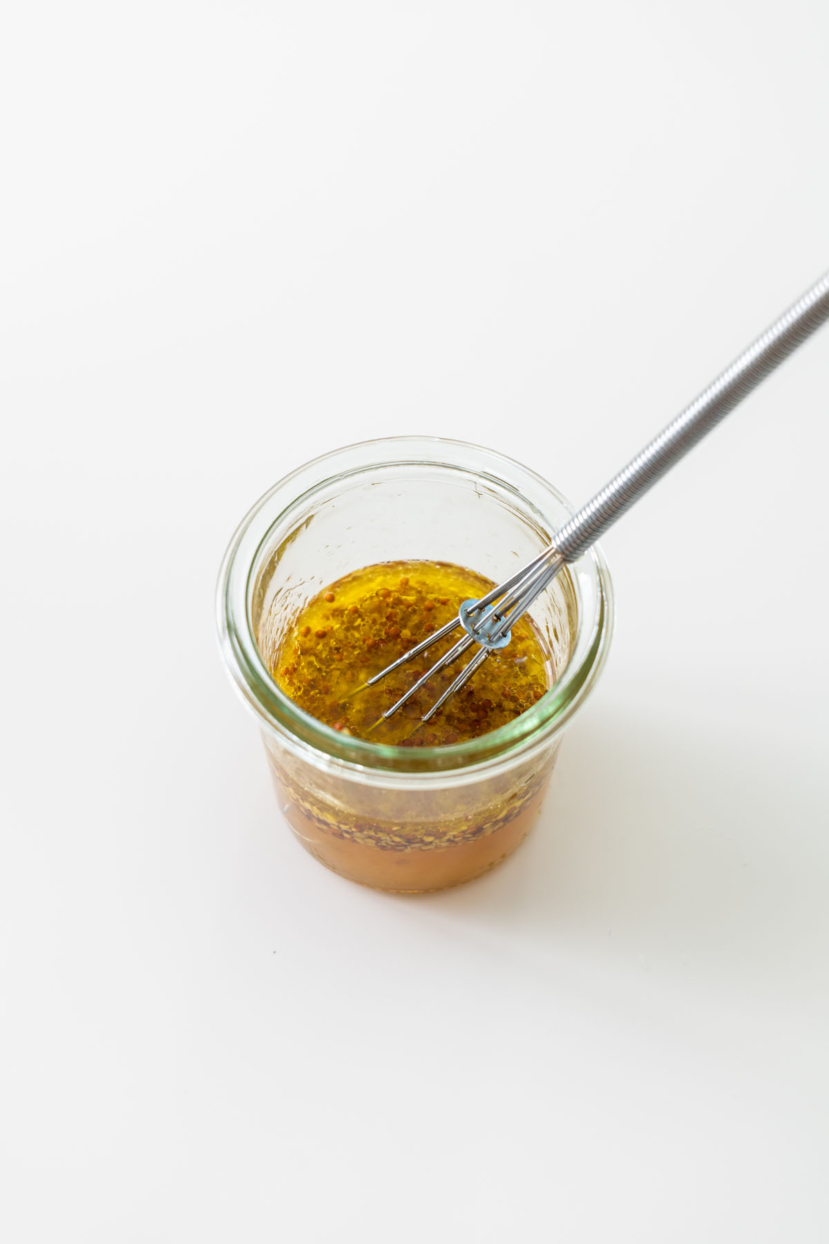Whole grain mustard dressing whisked in a small glass jar on a white surface.