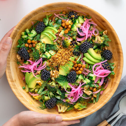 Crispy quinoa and blackberry salad in a wooden bowl being held, topped with avocado, cucumber, roasted chickpeas, dill, walnuts, and pickled onions.