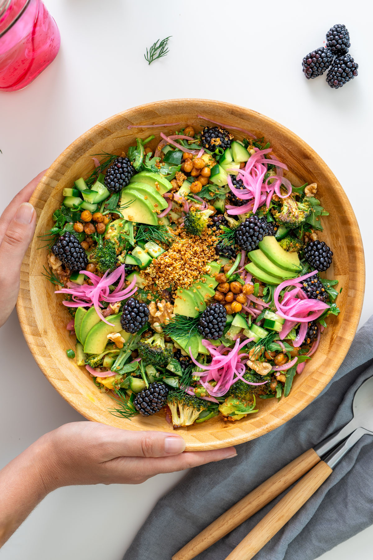 Crispy quinoa and blackberry salad in a wooden bowl being held, topped with avocado, cucumber, roasted chickpeas, dill, walnuts, and pickled onions.