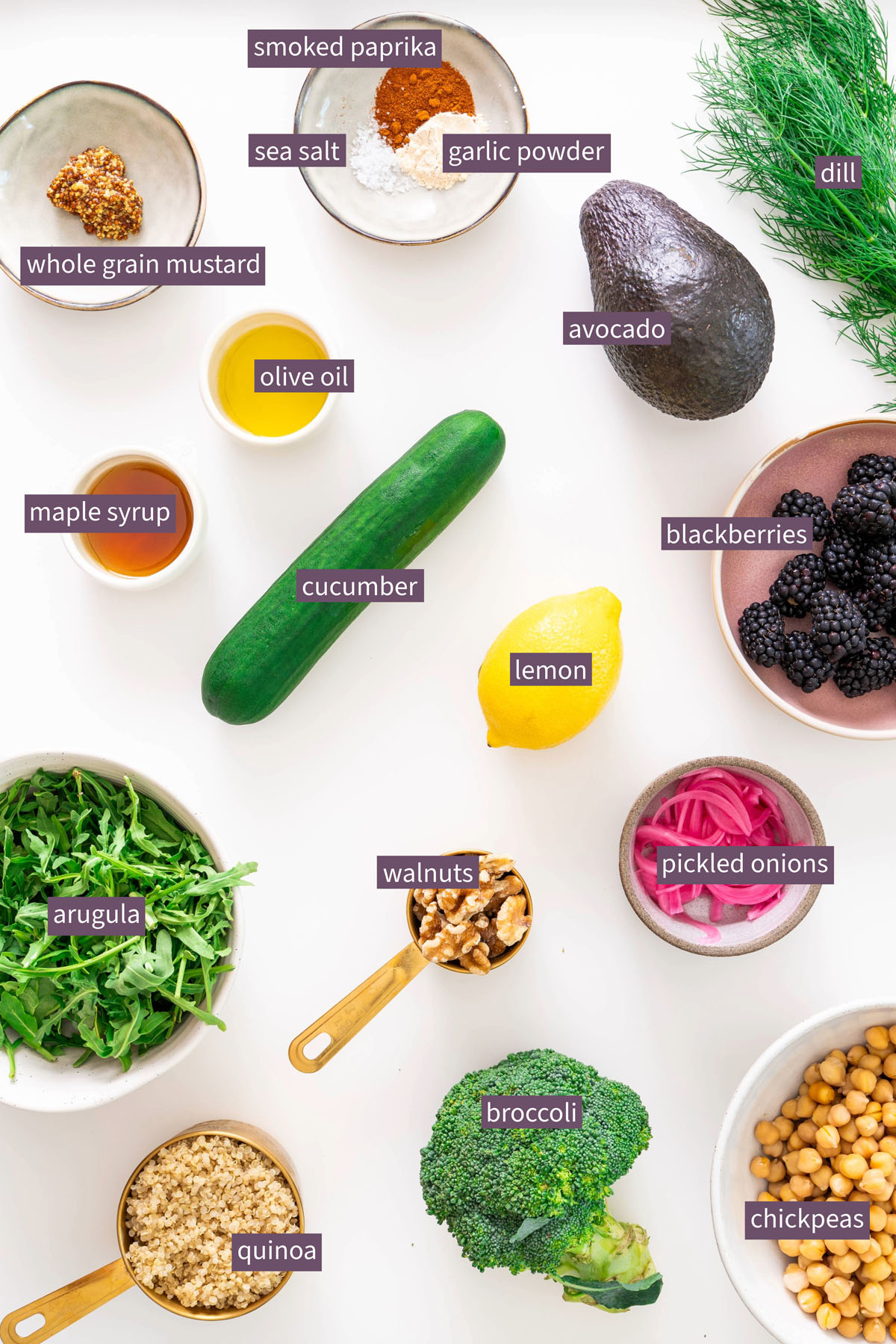 Labeled ingredients for crispy quinoa and blackberry salad arranged on a white surface, including blackberries, chickpeas, broccoli, quinoa, arugula, avocado, dill, and dressing ingredients.