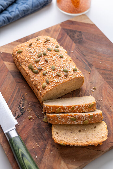 Red lentil bread loaf with three slices on a wooden cutting board with a bread knife, jar of red lentils in the background.