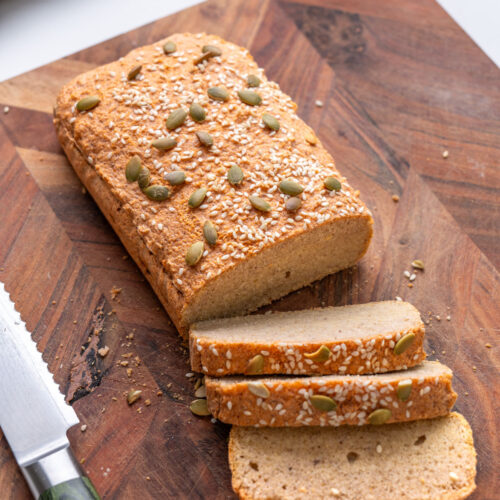 Red lentil bread loaf with three slices on a wooden cutting board with a bread knife, jar of red lentils in the background.