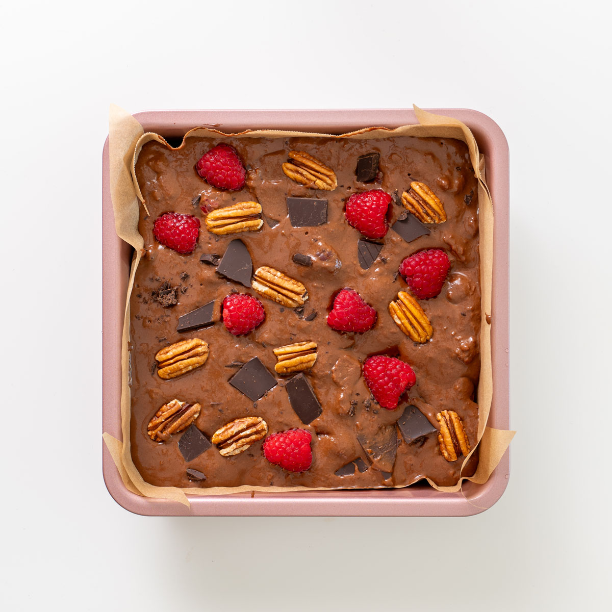 Unbaked brownie batter in a lined square pan topped with dark chocolate, pecans, and raspberries before baking.