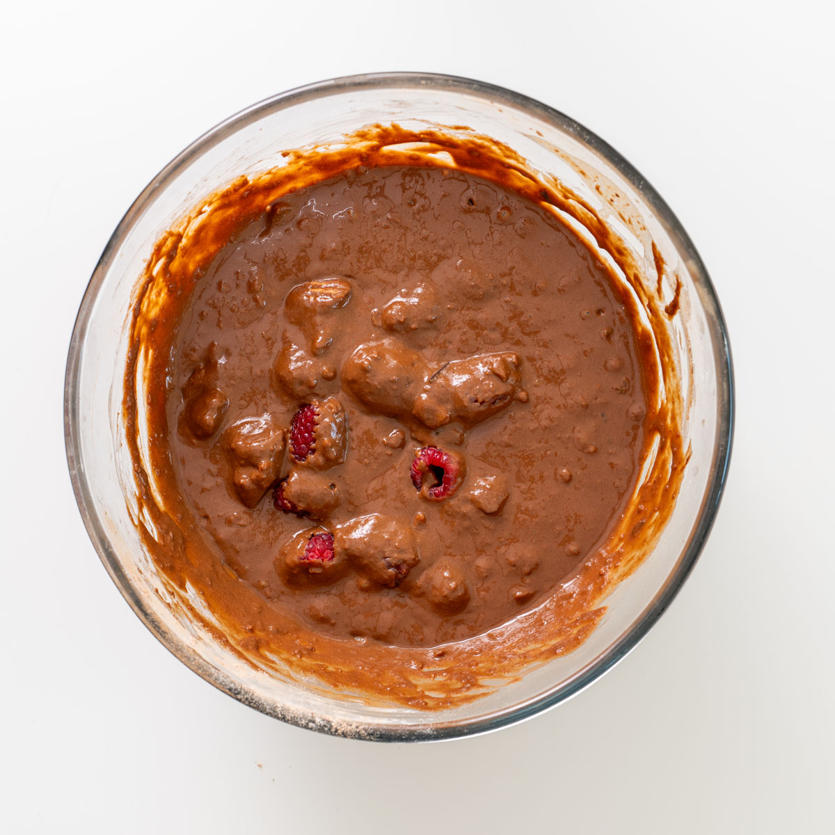Chocolate brownie batter with raspberries folded in, in a glass mixing bowl.