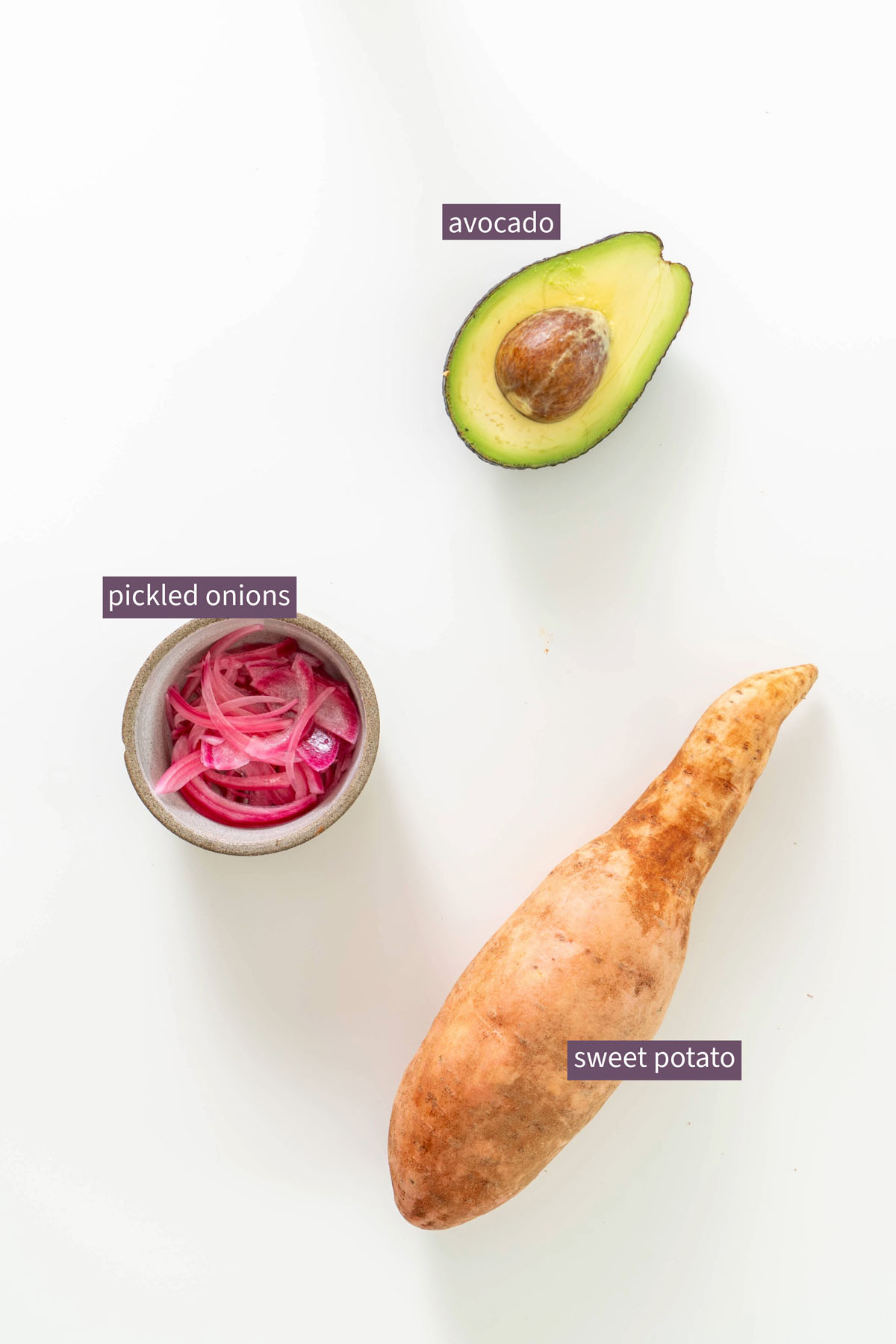 Flat lay of an avocado, pickled onions, and a whole sweet potato on a white background for a high-protein chipotle sweet potato bowl.