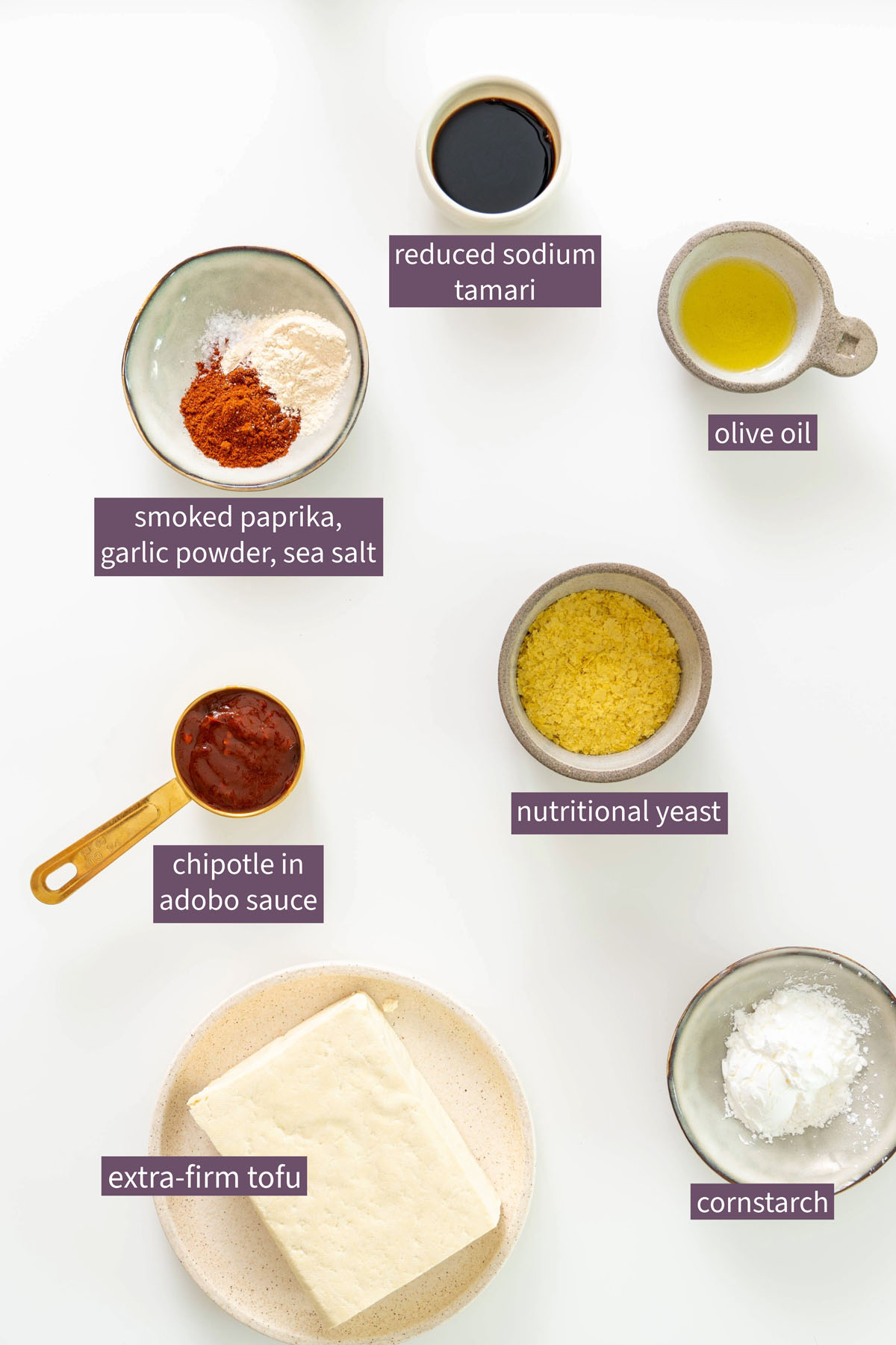 Flat lay of extra-firm tofu, chipotle in adobo sauce, nutritional yeast, cornstarch, reduced-sodium tamari, olive oil, smoked paprika, garlic powder, and sea salt on a white background.