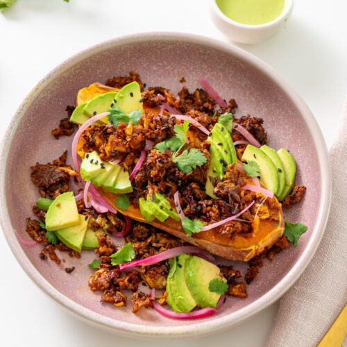 High-protein chipotle sweet potato bowl in a pink bowl with avocado, pickled onions, cilantro, and a small bowl of creamy cilantro jalapeño sauce.