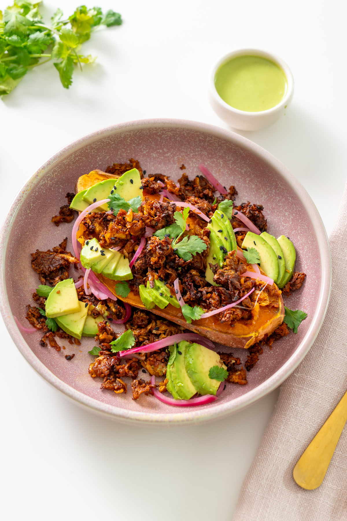 High-protein chipotle sweet potato bowl in a pink bowl with avocado, pickled onions, cilantro, and a small bowl of creamy cilantro jalapeño sauce.