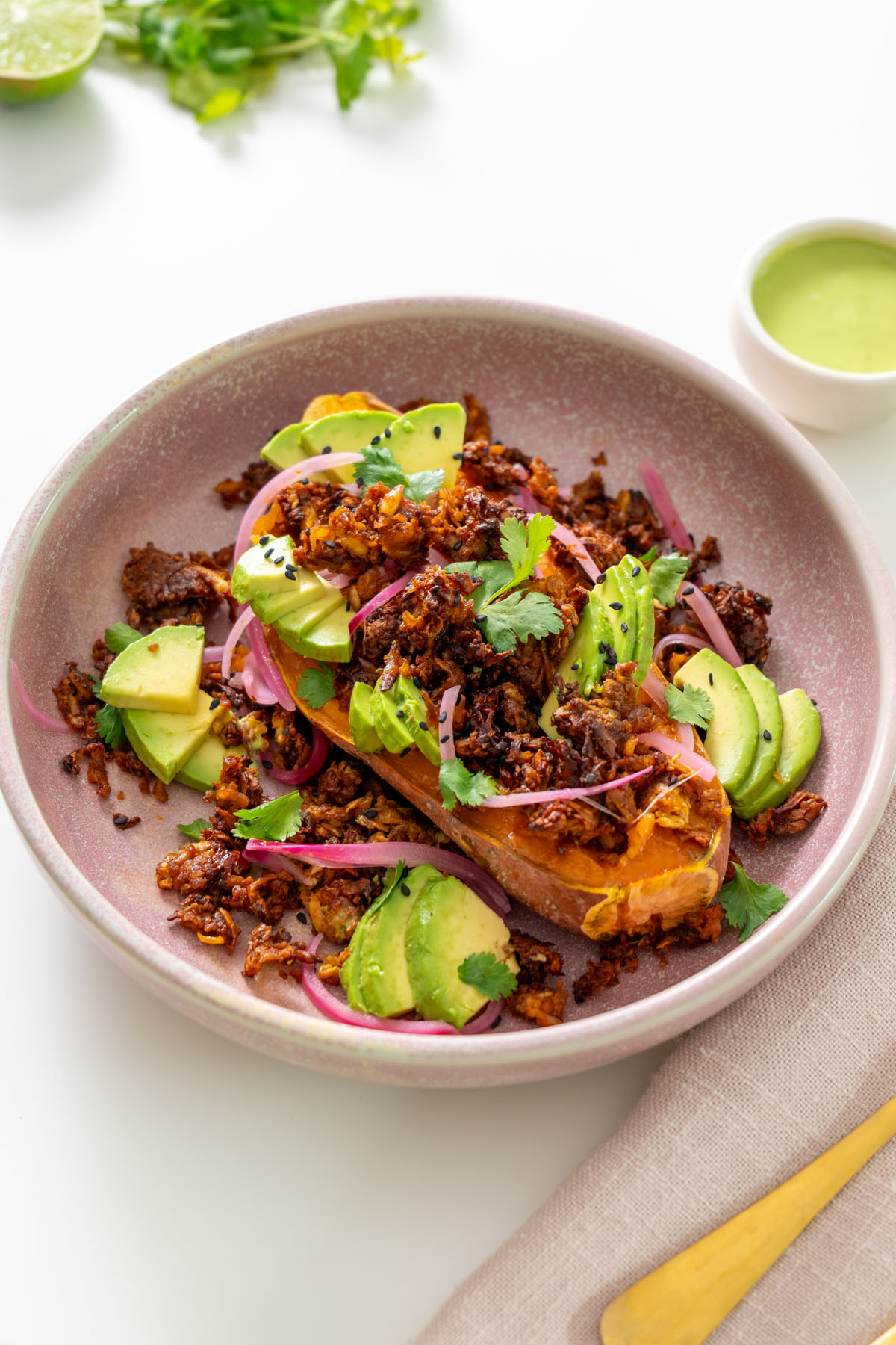 Close-up of a high-protein chipotle sweet potato bowl topped with avocado, pickled onions, cilantro, and crispy chipotle tofu, with green sauce on the side.