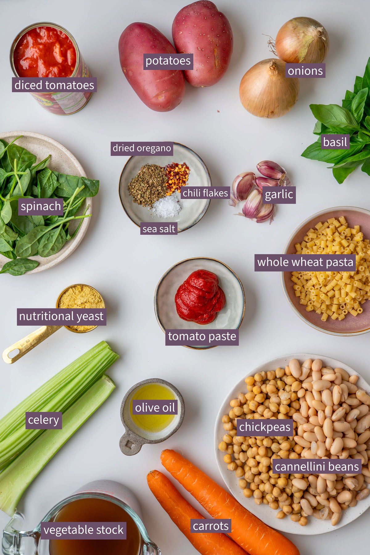 Vibrant ingredients for a longevity minestrone arranged on a white surface, including potatoes, onions, carrots, celery, chickpeas, cannellini beans, tomato paste, spinach, basil, and whole wheat pasta