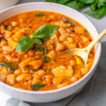 A bowl of longevity minestrone with cannellini beans, chickpeas, potatoes, and spinach in a rich tomato broth, garnished with fresh basil and a gold spoon