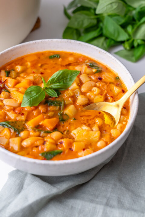 A bowl of longevity minestrone with cannellini beans, chickpeas, potatoes, and spinach in a rich tomato broth, garnished with fresh basil and a gold spoon