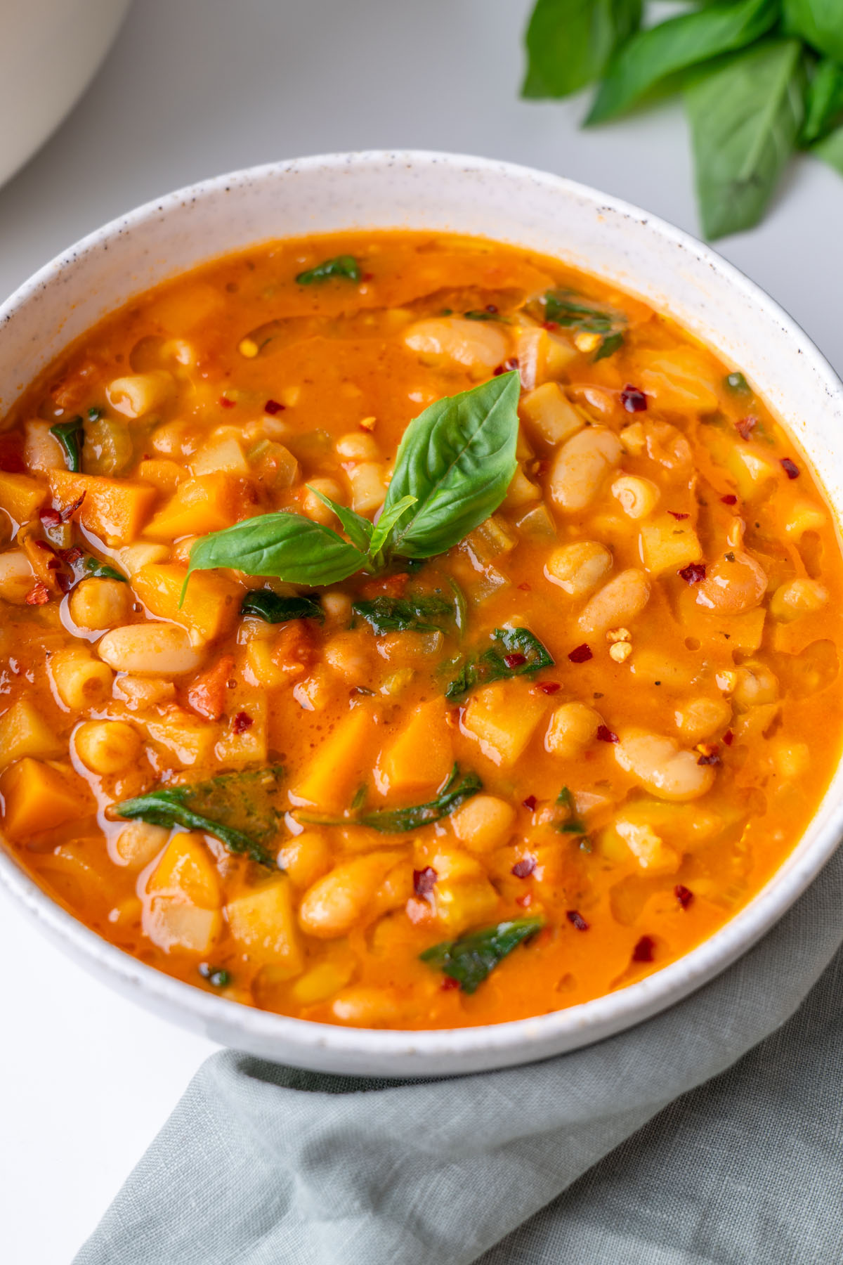 Close-up of longevity minestrone showing cannellini beans, chickpeas, potatoes, wilted spinach, and chili flakes in a rich tomato broth, topped with fresh basil