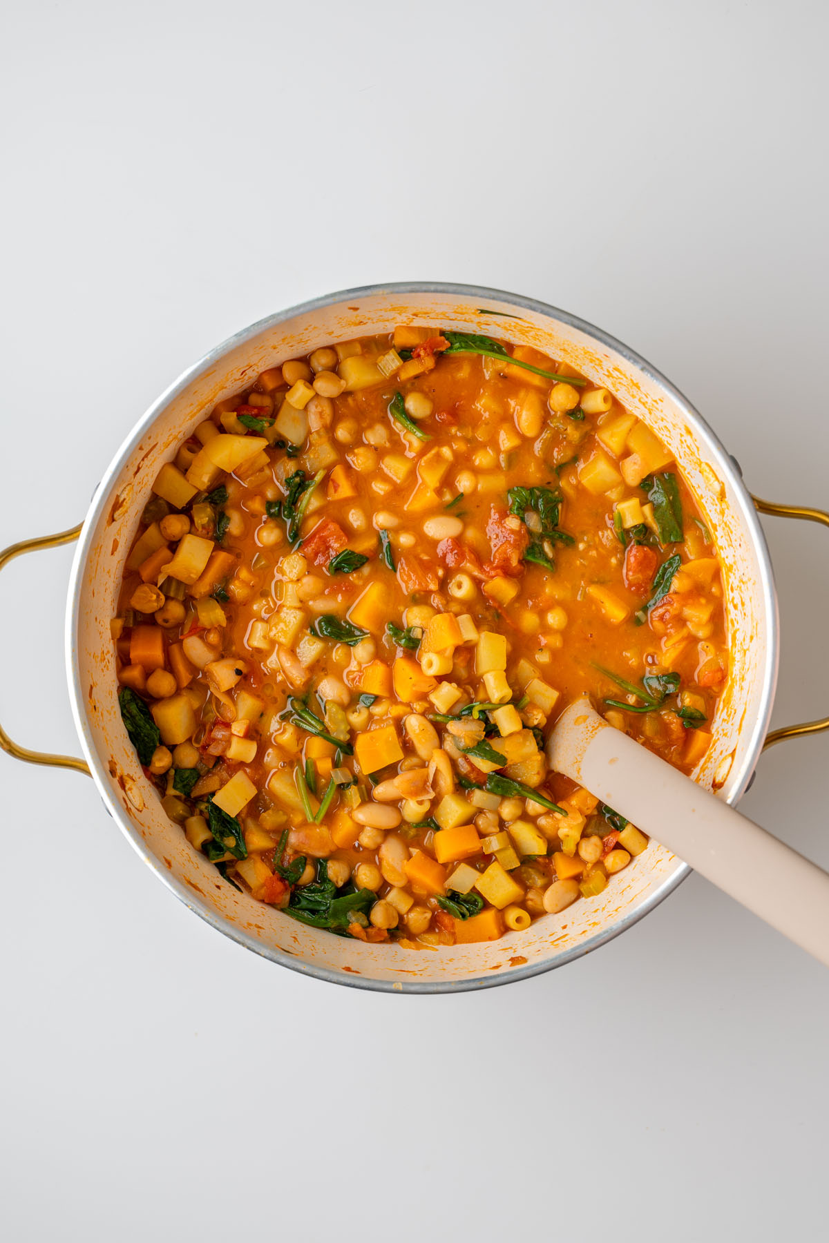 Overhead view of a large pot of longevity minestrone with a spatula, showing beans, chickpeas, pasta, spinach, and vegetables in a tomato broth