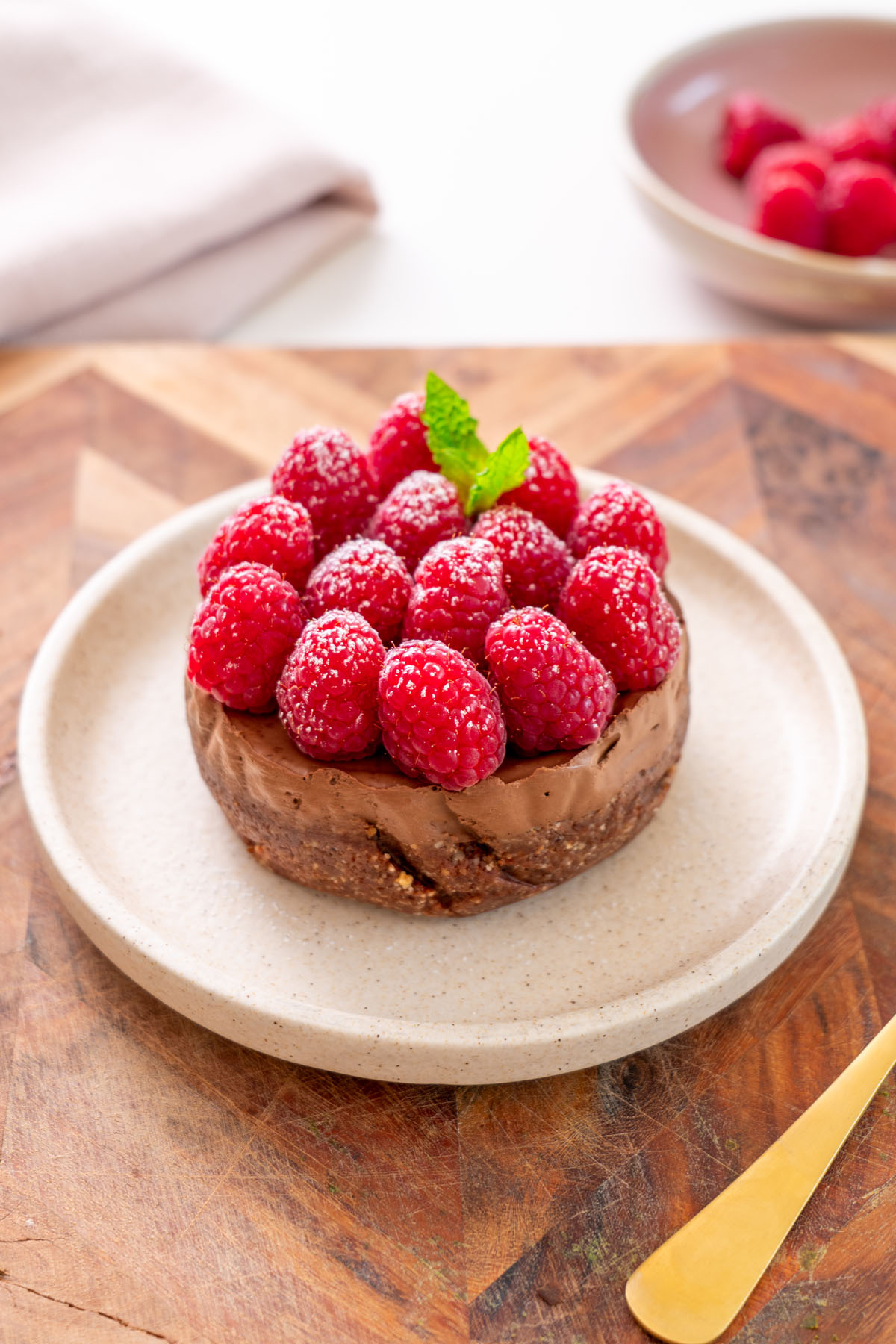 No-bake mini chocolate tart topped with fresh raspberries on a plate on a wooden board, with a gold spoon nearby.