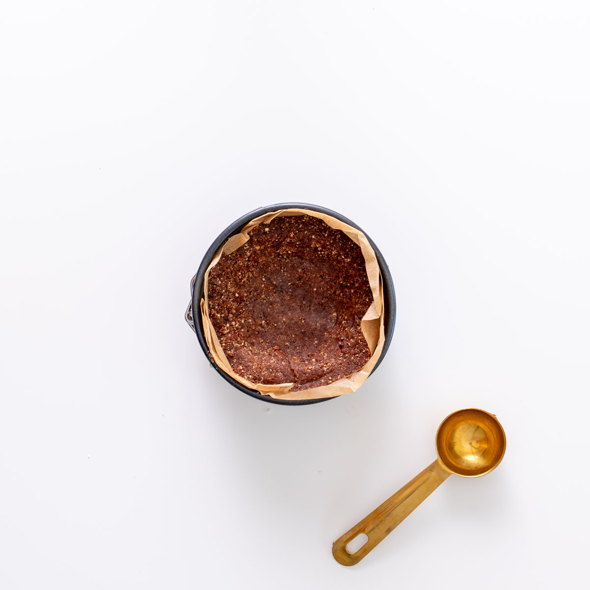 Hazelnut cacao crust pressed into a parchment-lined mini tart pan on a white background, with a gold measuring spoon beside it.