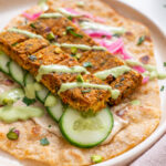 Close-up of sheet pan tofu kebab pieces on a flatbread with cucumber slices, pickled pink onions, creamy green sauce drizzle, and hummus