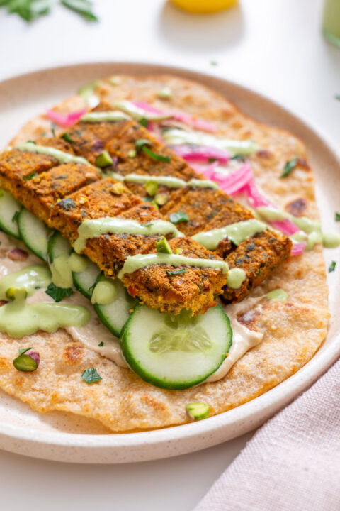 Close-up of sheet pan tofu kebab pieces on a flatbread with cucumber slices, pickled pink onions, creamy green sauce drizzle, and hummus