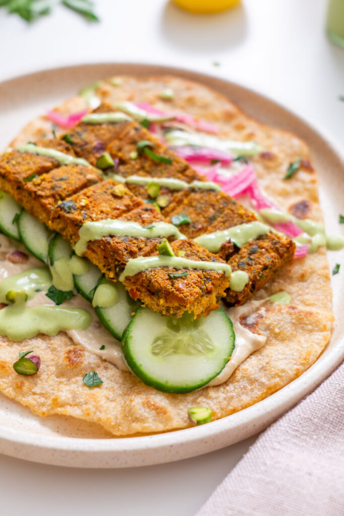 Close-up of sheet pan tofu kebab pieces on a flatbread with cucumber slices, pickled pink onions, creamy green sauce drizzle, and hummus