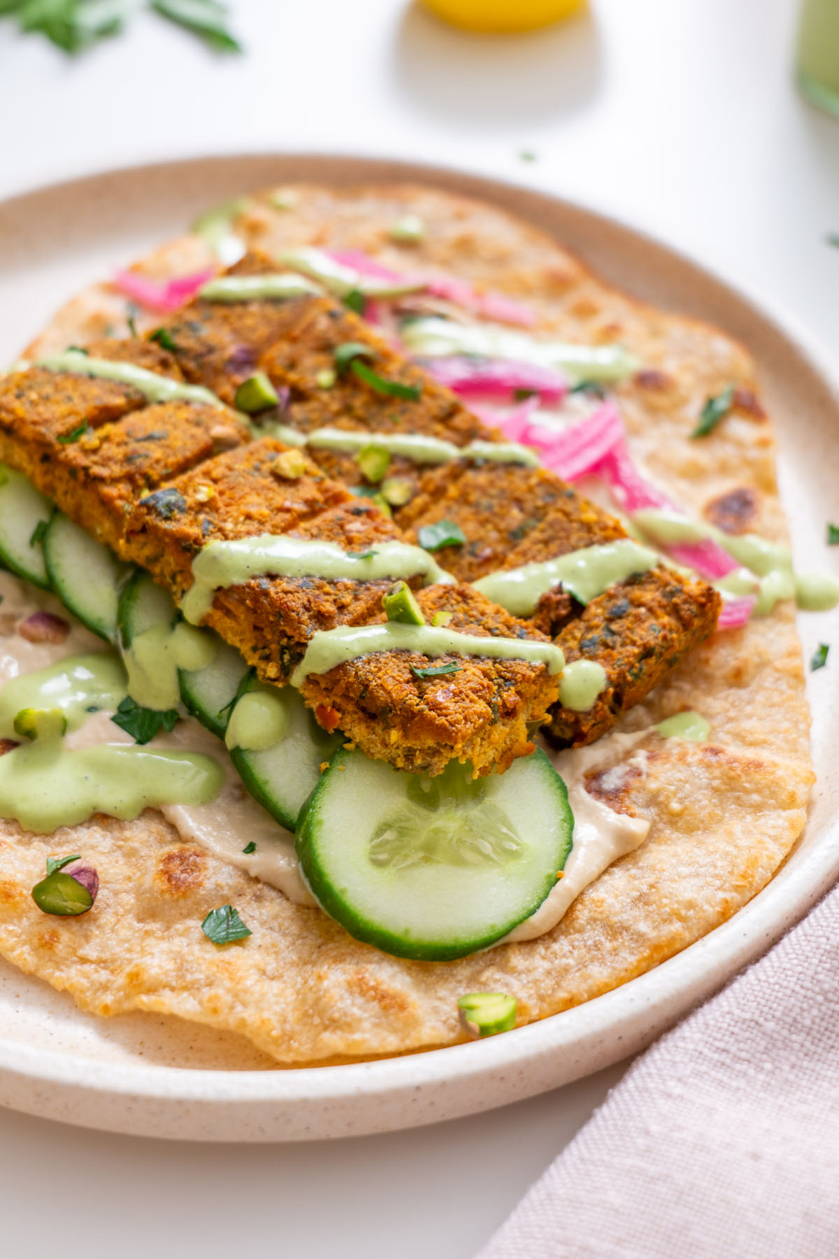 Close-up of sheet pan tofu kebab pieces on a flatbread with cucumber slices, pickled pink onions, creamy green sauce drizzle, and hummus