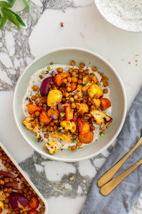 Styled overhead shot of a sweet and spicy Mediterranean bowl on a marble surface, with a sheet pan of roasted chickpeas and veggies, a bowl of mint yogurt sauce, fresh mint sprigs, gold utensils, and a blue linen napkin.