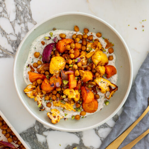 Styled overhead shot of a sweet and spicy Mediterranean bowl on a marble surface, with a sheet pan of roasted chickpeas and veggies, a bowl of mint yogurt sauce, fresh mint sprigs, gold utensils, and a blue linen napkin.