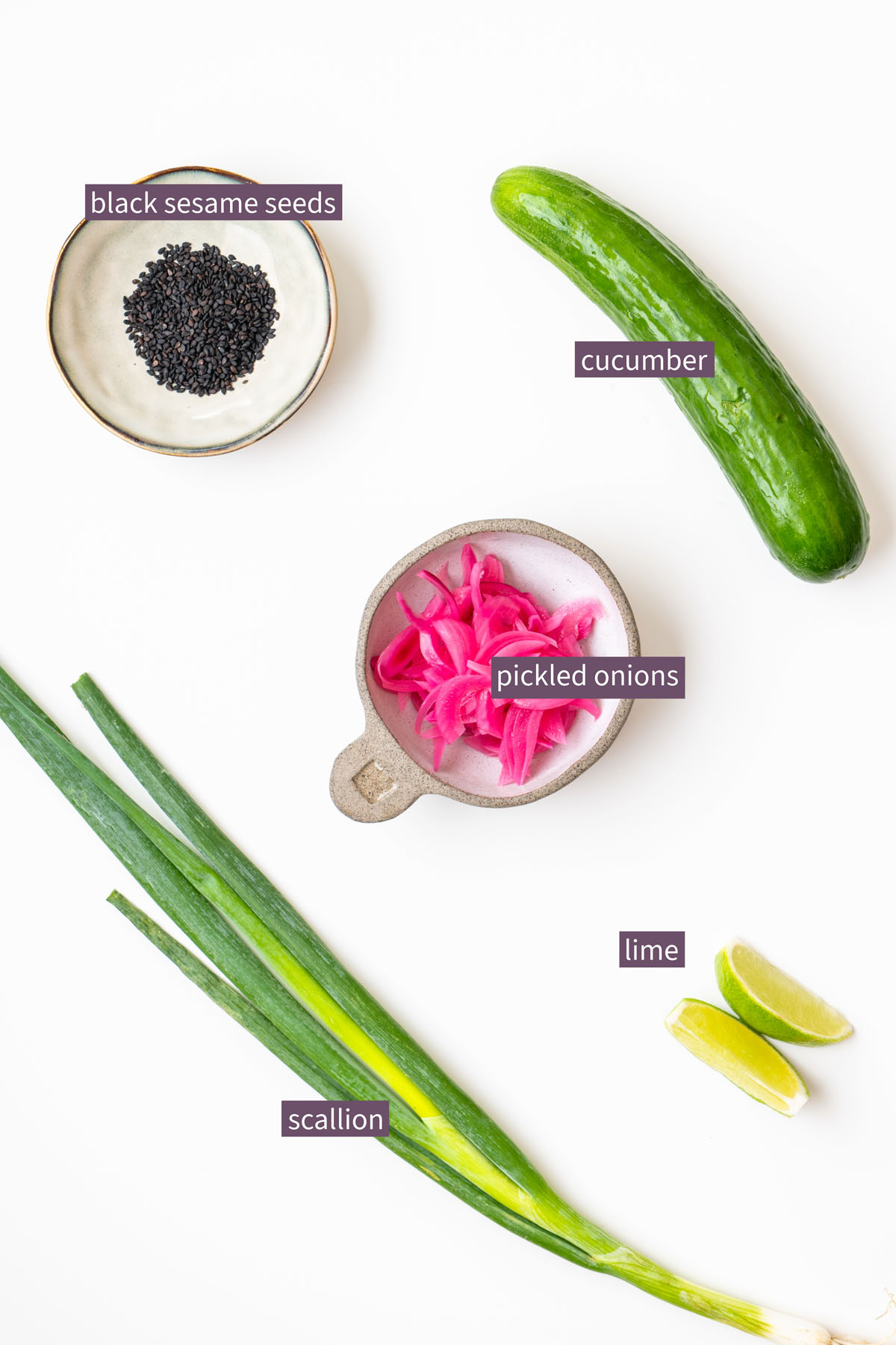 Vibrant topping ingredients for a gochujang broccoli tofu bowl on a white surface, including black sesame seeds, cucumber, pickled onions, scallion, and lime wedges