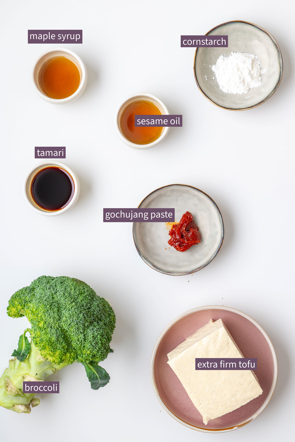 Vibrant ingredients for sticky gochujang tofu on a white surface, including maple syrup, cornstarch, sesame oil, tamari, gochujang paste, broccoli, and extra firm tofu