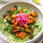 Close-up of a gochujang broccoli tofu bowl with sticky golden tofu, charred broccoli, pickled onions, cucumber, scallions, and black sesame seeds over creamy edamame avocado spread