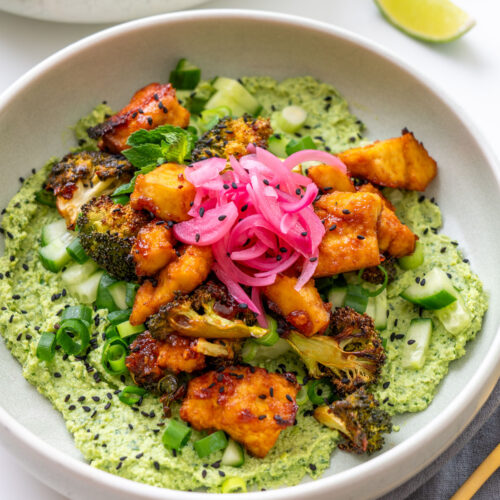Close-up of a gochujang broccoli tofu bowl with sticky golden tofu, charred broccoli, pickled onions, cucumber, scallions, and black sesame seeds over creamy edamame avocado spread