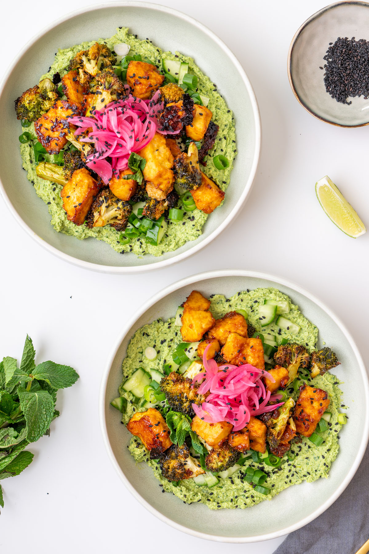 Two gochujang broccoli tofu bowls with sticky tofu, roasted broccoli, pickled onions, cucumber, scallions, and black sesame seeds over creamy edamame avocado spread, with fresh mint and lime on the side