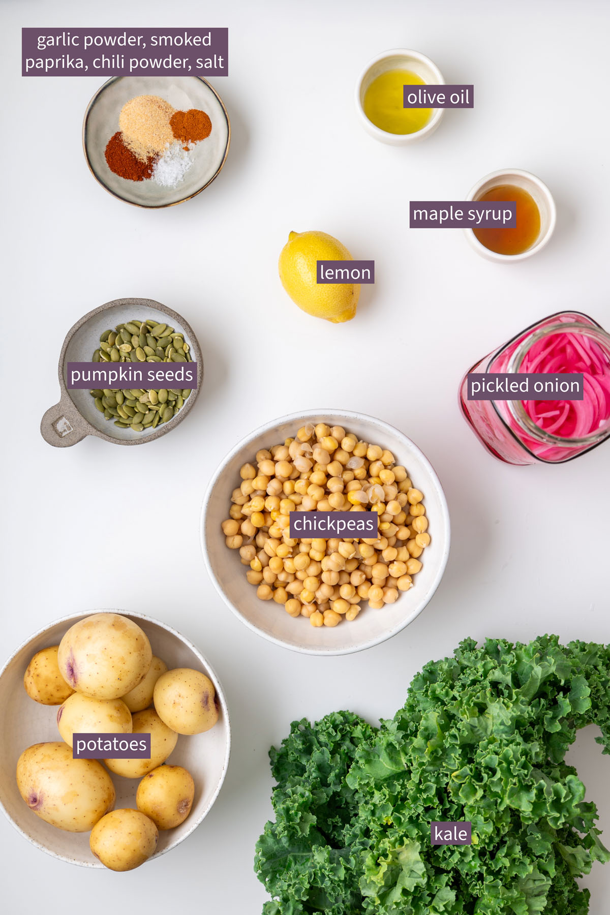 Vibrant ingredients for a roasted potato and avocado ranch salad on a white surface, including potatoes, chickpeas, kale, pumpkin seeds, spices, olive oil, maple syrup, lemon, and pickled onion