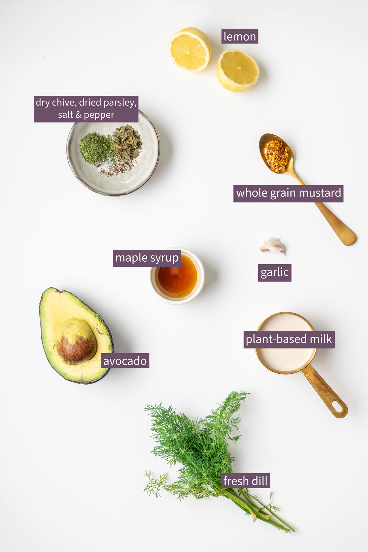 Vibrant ingredients for an avocado ranch dressing on a white surface, including avocado, fresh dill, lemon, plant-based milk, whole grain mustard, maple syrup, garlic, and dried herbs