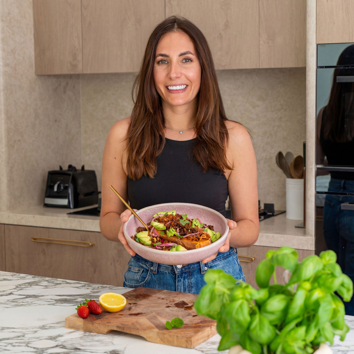 Sarah Cobacho, Plantbaes founder, smiling in her kitchen holding a vibrant plant-based bowl