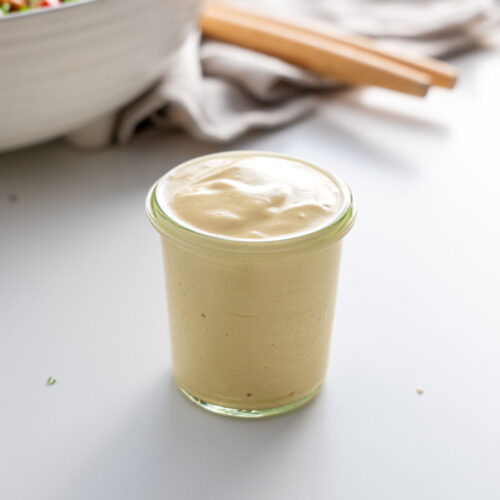 Jar of creamy cashew Caesar dressing on a white surface with a salad bowl in the background.